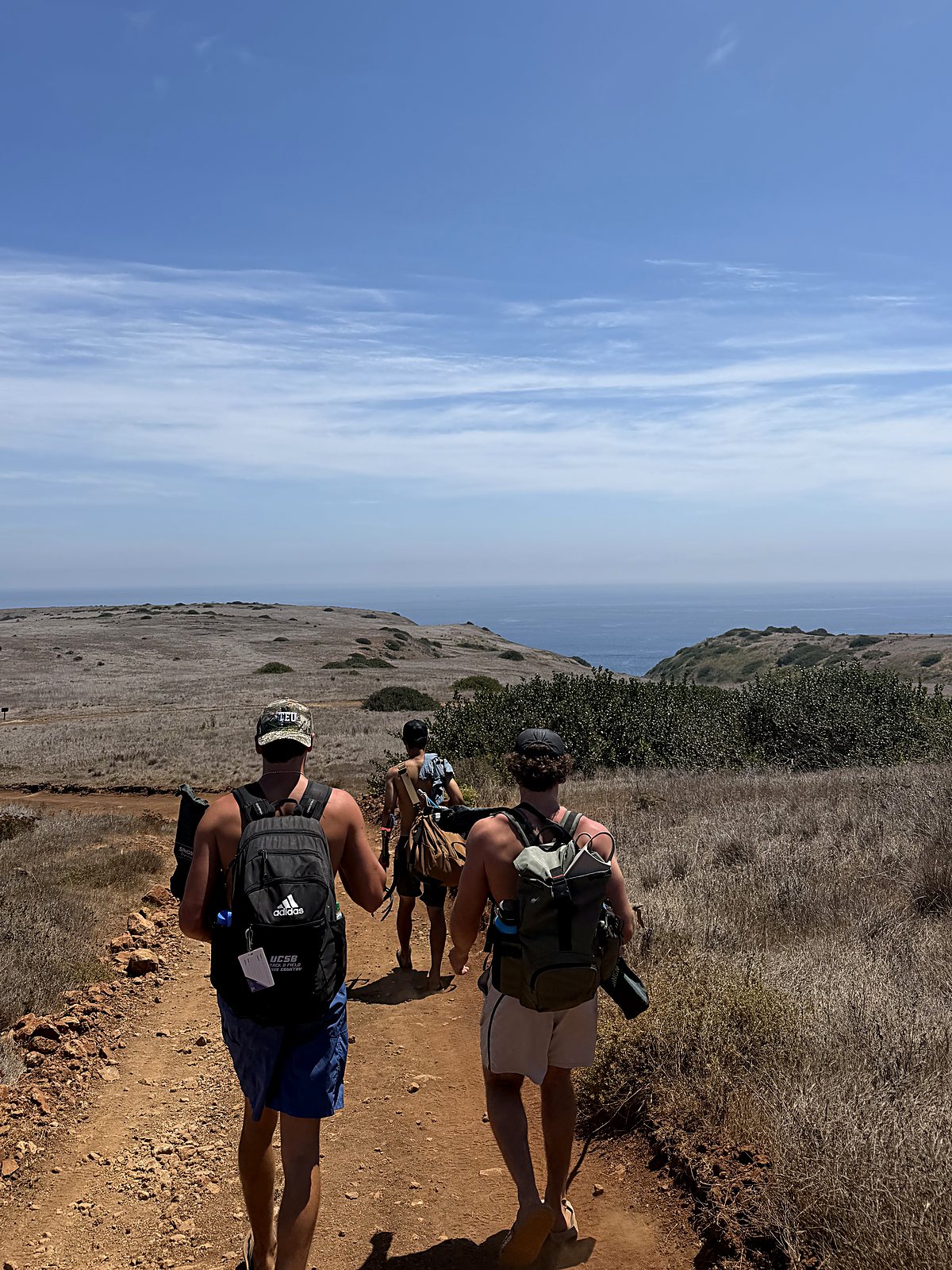 Three of the Lavare crew hiking a dirt road across Santa Cruz Island with packs and dive gear, the Pacific ahead of them.