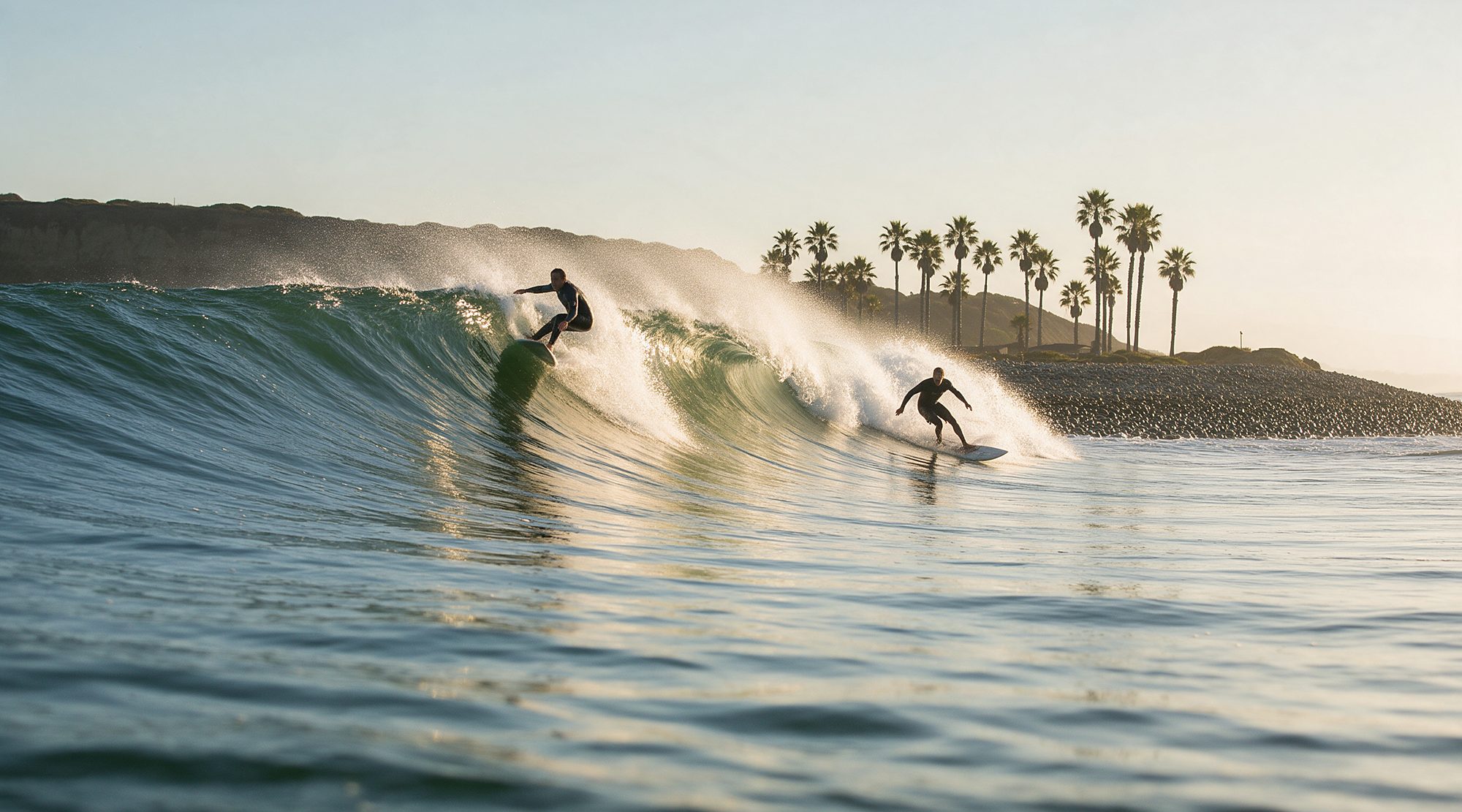 Two surfers riding the same glassy right-hand wave peeling down the point at Rincon, palms along the bluff behind them in early morning light.