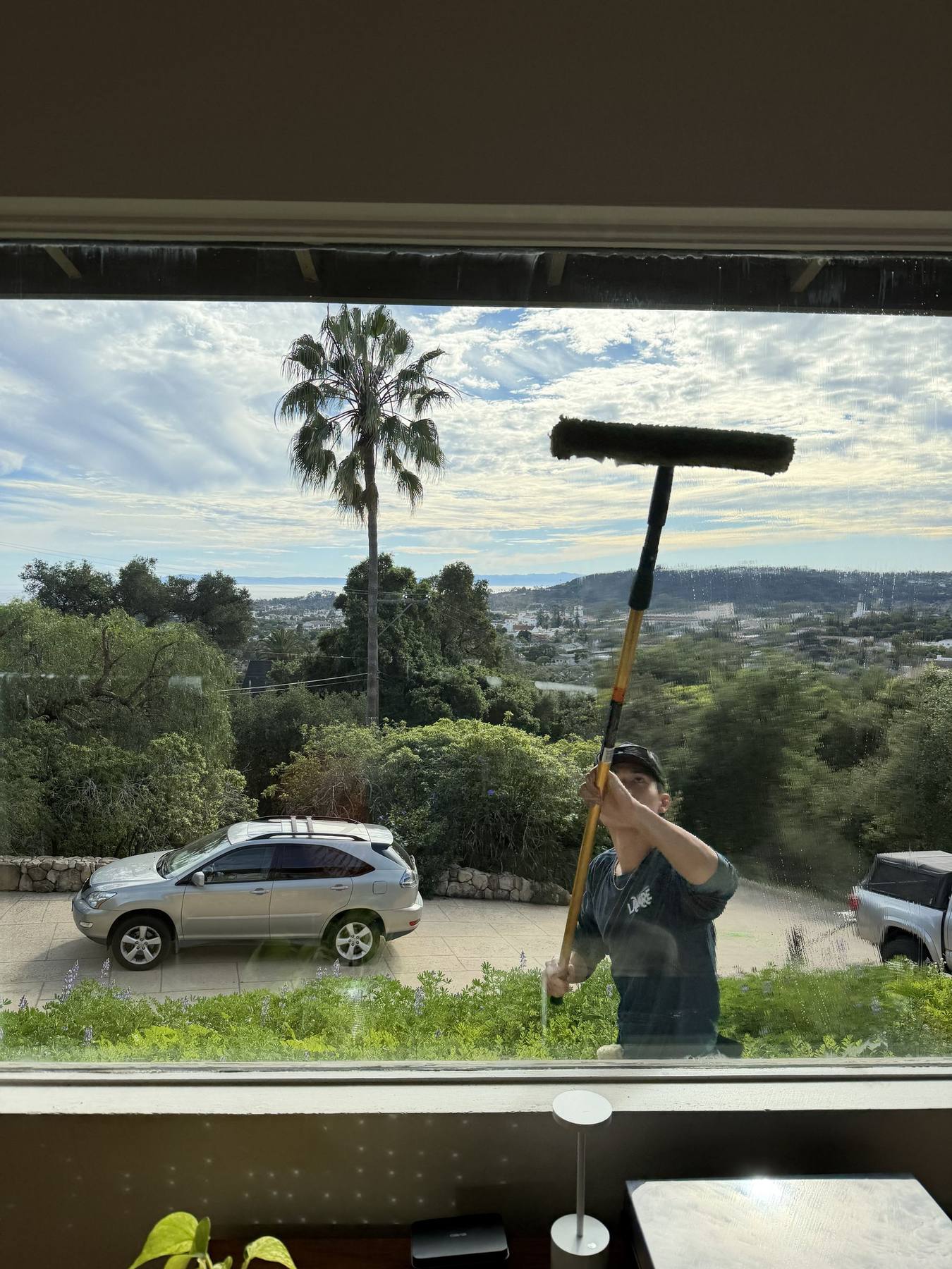 A Lavare technician reaching a second-story window with a water-fed pole, palms and ocean haze behind the house.